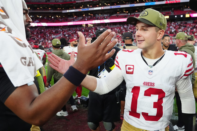 A football player in a white 49ers jersey (#13) and cap reaches out to high-five a teammate wearing a towel on his shoulder in a crowded stadium.